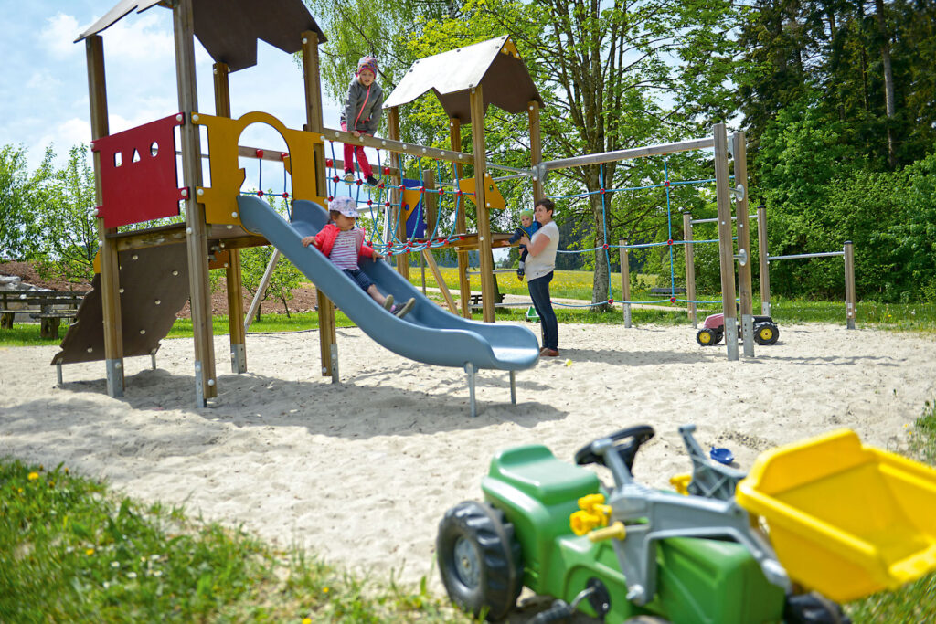 Der Spielplatz der Sonne im Aischfeld. Ein Kind spielt auf dem Klettergerüst, ein weiteres Kind rutscht die Rutsche hinunter. Eine Mutter mit einem weiteren Kind im Arm steht daneben