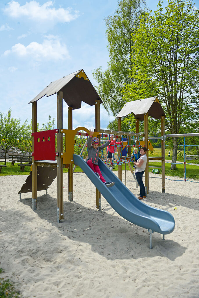 Der Spielplatz der Sonne im Aischfeld. Ein Kind spielt auf dem Klettergerüst, ein weiteres Kind rutscht die Rutsche hinunter. Eine Mutter mit einem weiteren Kind im Arm steht daneben