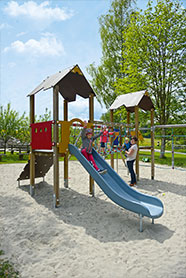 Der Spielplatz der Sonne im Aischfeld. Ein Kind spielt auf dem Klettergerüst, ein weiteres Kind rutscht die Rutsche hinunter. Eine Mutter mit einem weiteren Kind im Arm steht daneben